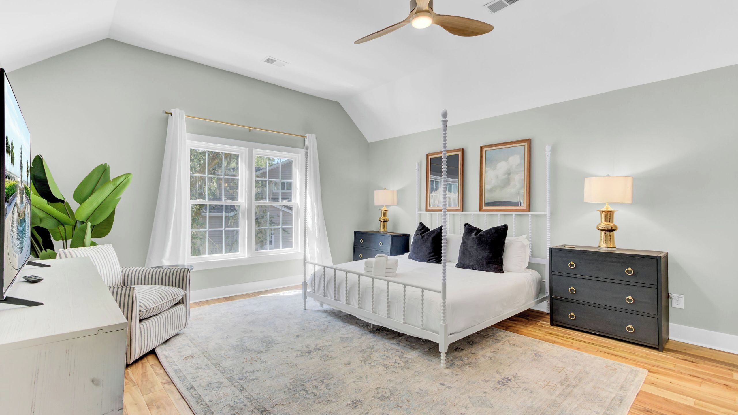 A bedroom with a bed, bedside tables, a desk, a chair, a large window, and a ceiling fan.