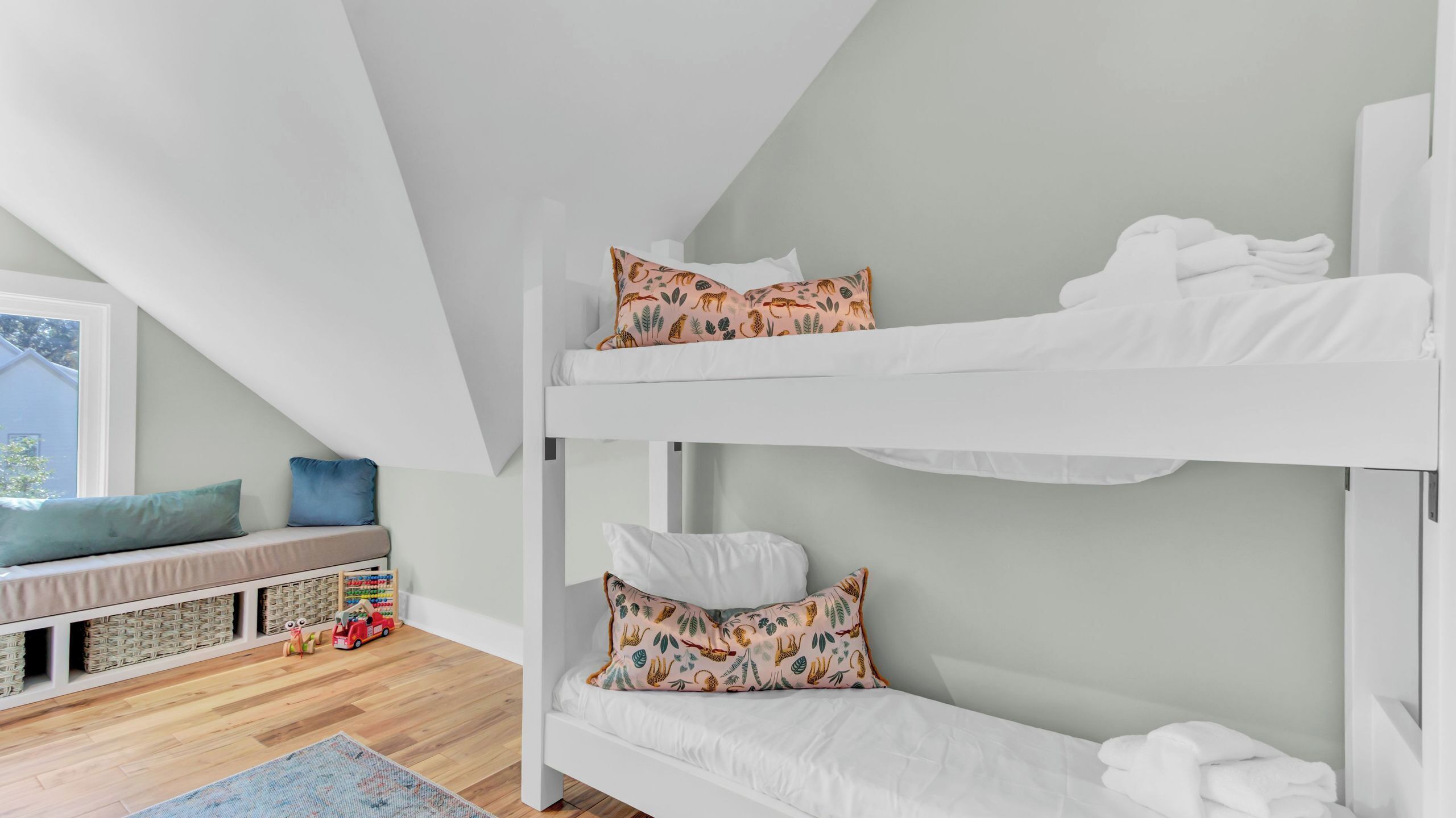 A bunk bed with white bedding, a window with a view, a small seating area with cushions, and a wooden floor.
