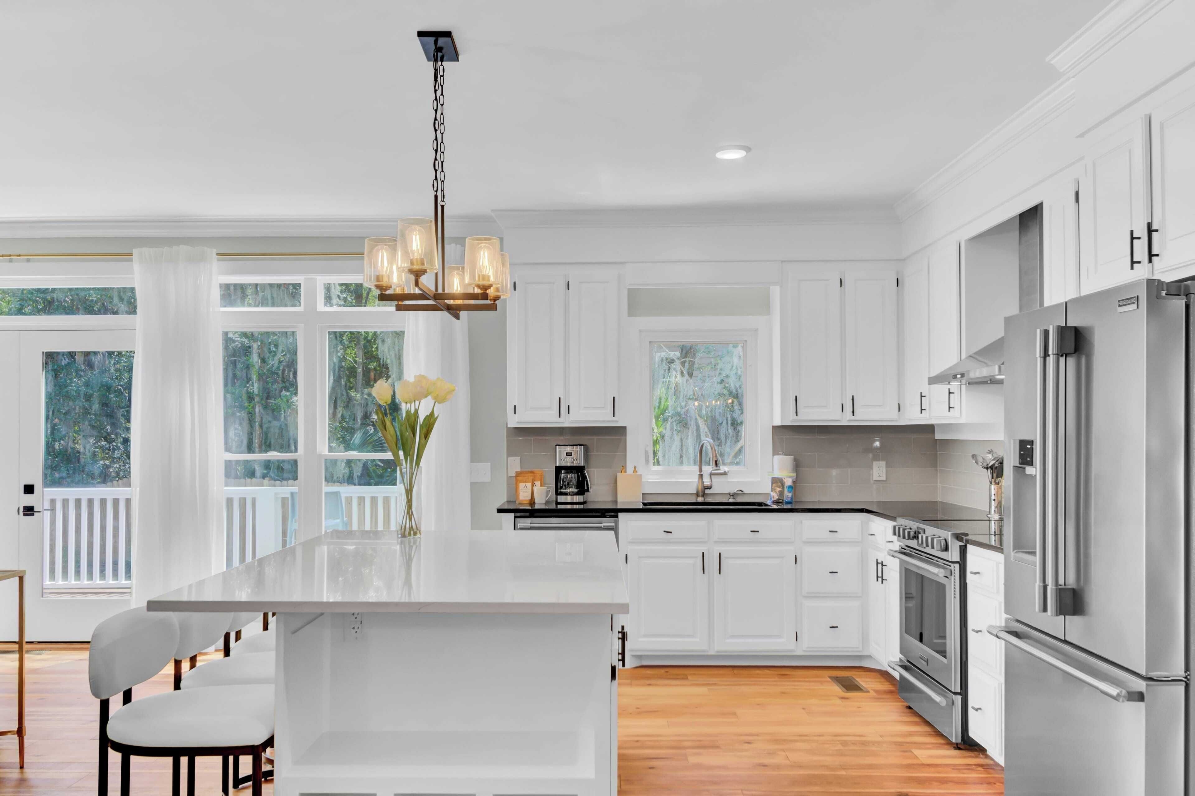 A modern kitchen with white cabinetry, a large island, stainless steel appliances, and a view of the outdoors through large windows.