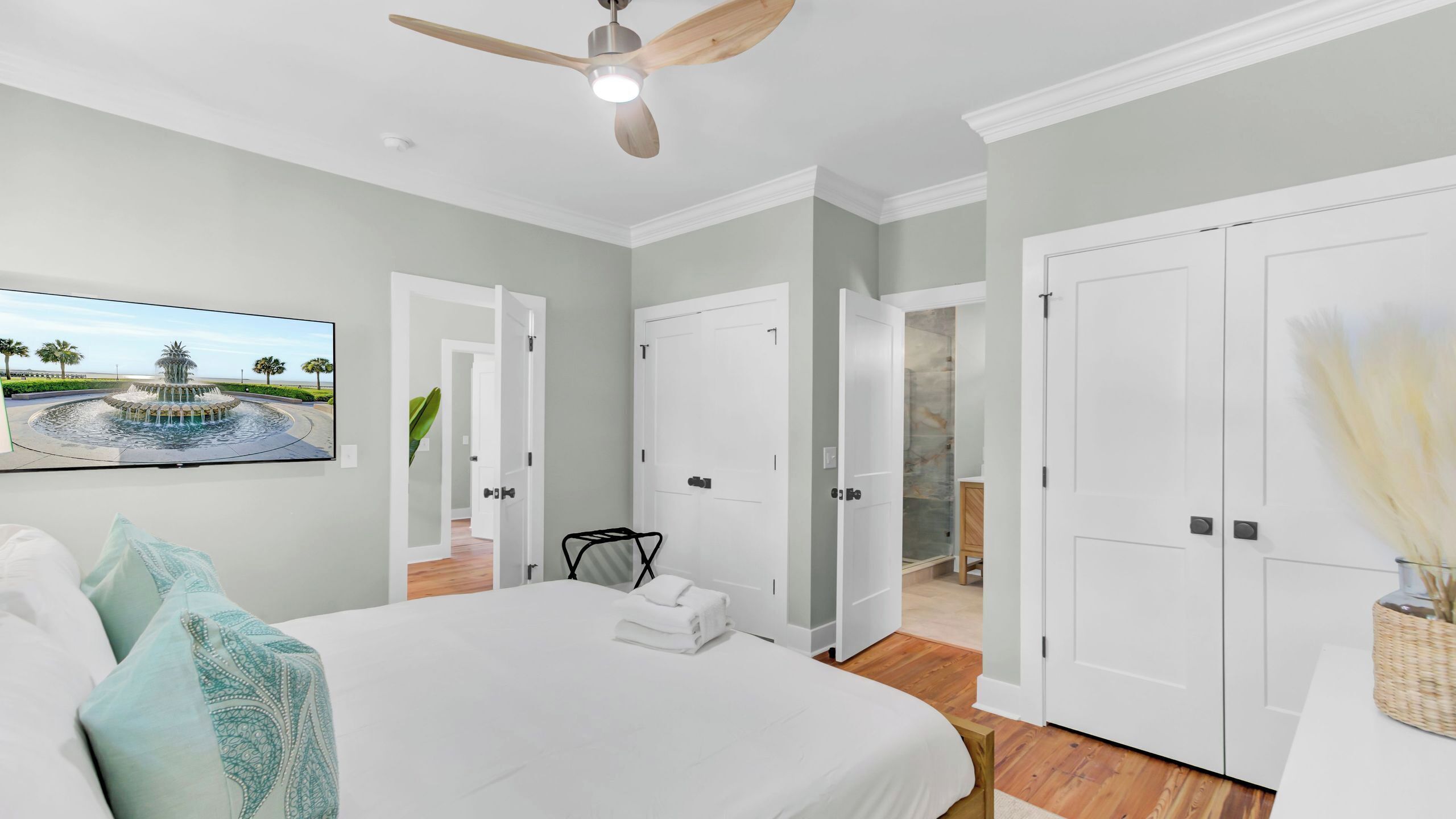 A bedroom with a bed, a ceiling fan, a painting of a fountain, and white doors leading to other rooms.