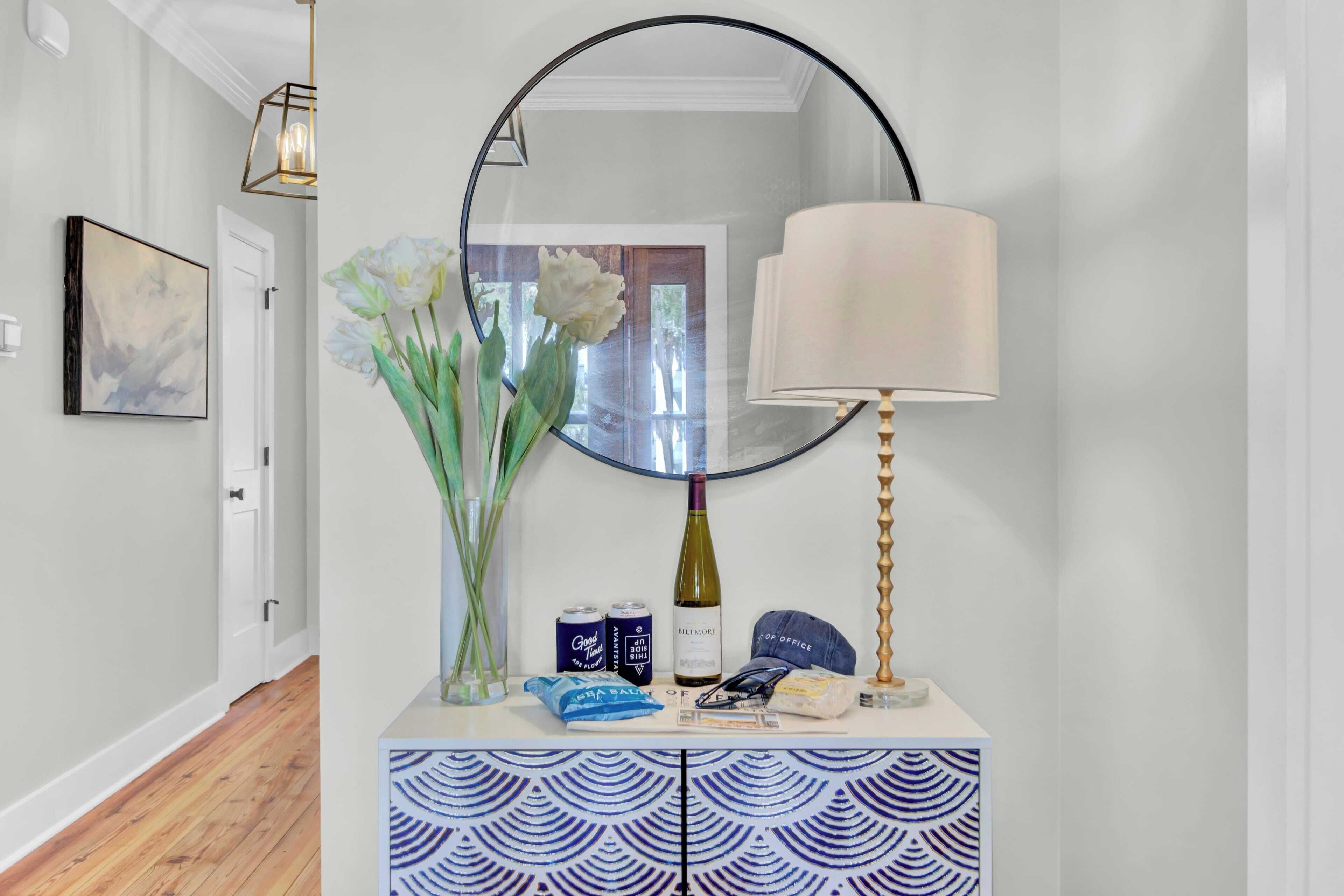 A hallway with a white door, a framed abstract painting, a vase with white flowers, a wine bottle, cans, a blue bag, a cap, and a patterned cabinet.