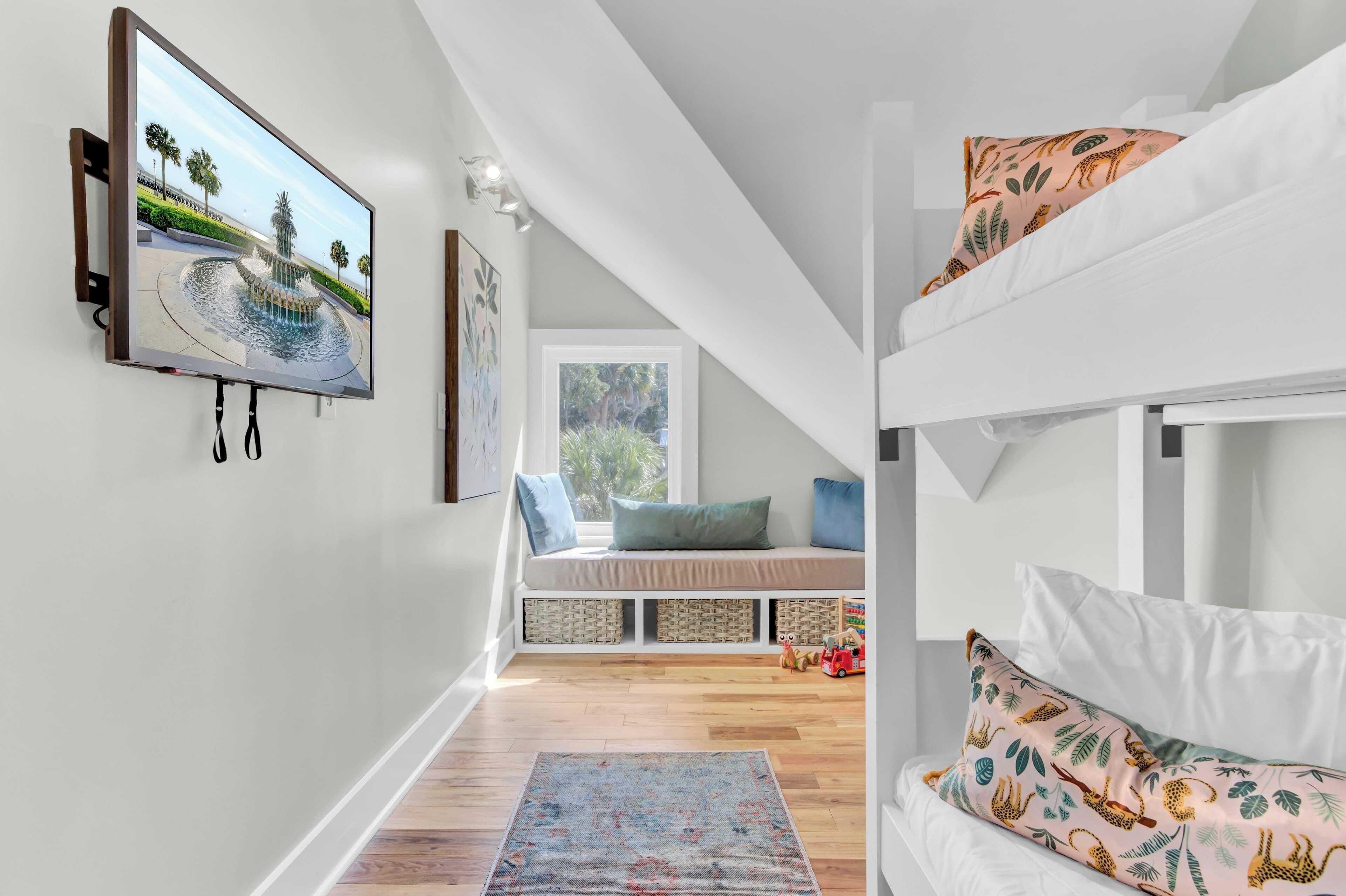 A room with a bunk bed, a flat-screen TV mounted on the wall, a window seat with cushions, and a colorful rug.