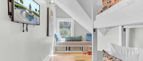 A room with a bunk bed, a flat-screen TV mounted on the wall, a window seat with cushions, and a colorful rug.