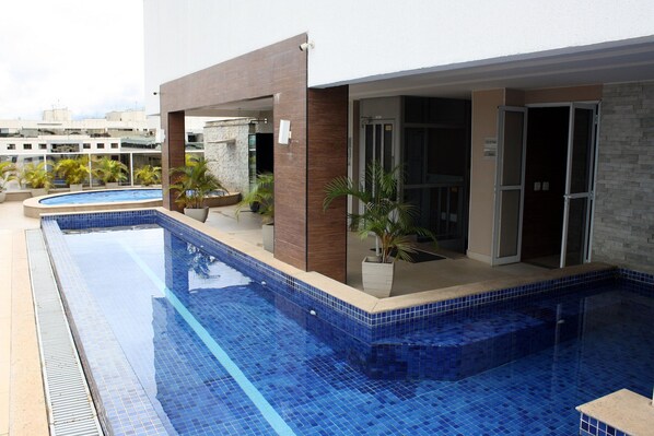 A swimming pool with a tiled edge, surrounded by a building with a modern design, including a stone wall and glass doors.