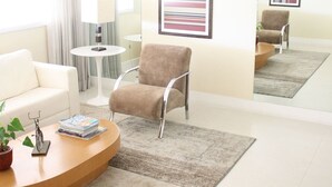 A modern hotel room with a beige sofa, a round wooden coffee table, a brown armchair, and a small side table with a lamp.