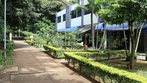 Un chemin bordé de haies et d’arbres soigneusement taillés, qui mène à un bâtiment moderne doté d’un auvent bleu.