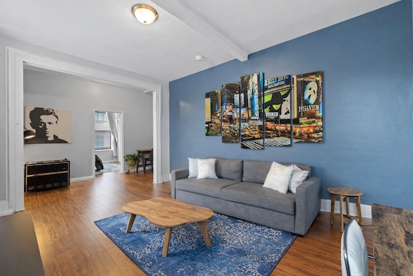 A living room with a gray sofa, a wooden coffee table, and a blue rug. There is a wall with a cityscape painting and a framed picture of a person.