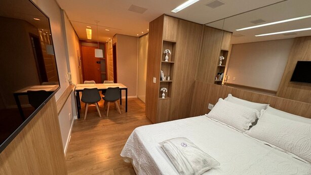 A hotel room with a bed, a desk, and chairs. There is a wooden wardrobe and a shelf with decorative items. - Flat Sudoeste Ed. Leonardo (Brasília)