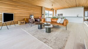 Un salon moderne avec un plafond en bois, un grand téléviseur posé sur un meuble, un canapé en cuir, une table basse et un coin cuisine avec des meubles blancs.