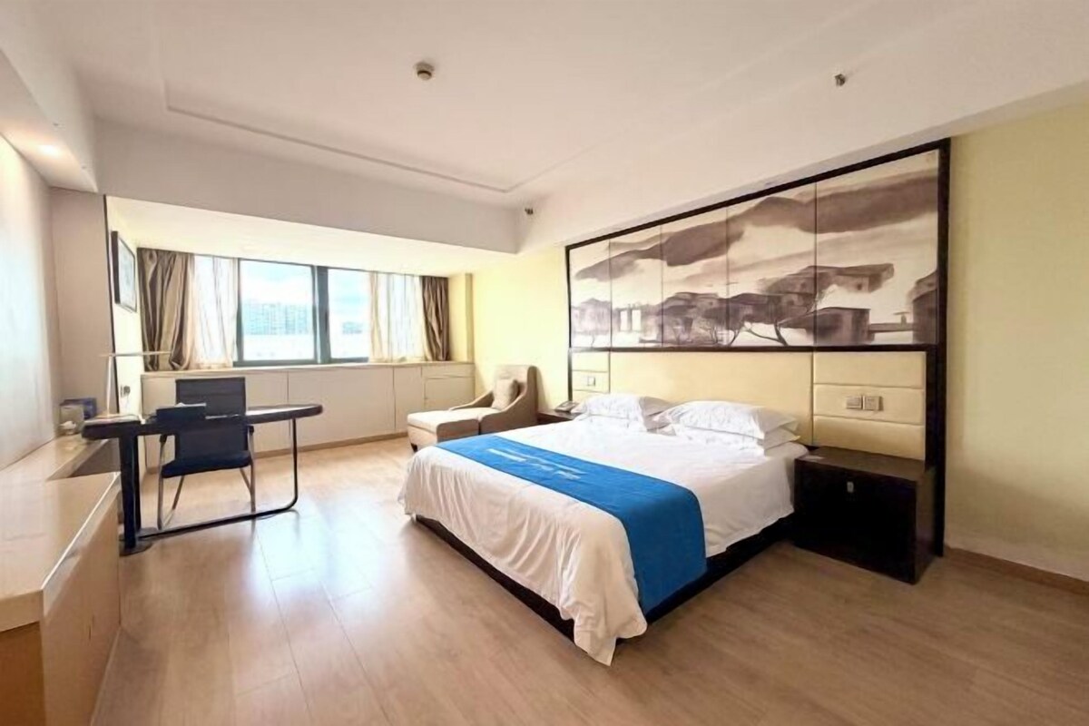 Ett modernt hotellrum med en stor säng, ett skrivbord och en stol. Det finns ett fönster med gardiner, en tavla på väggen och utsikt över staden.