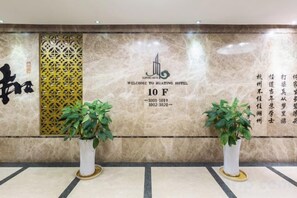 A hotel reception area with a marble wall, a welcome sign, and two potted plants. - Huating Hotel (Huzhou)
