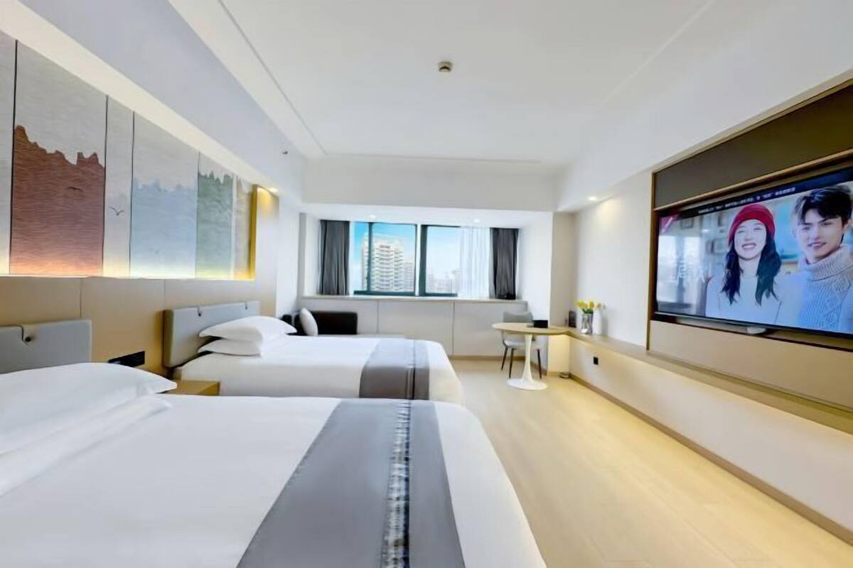 A hotel room with two beds, a TV, a small table, and a window with a city view.