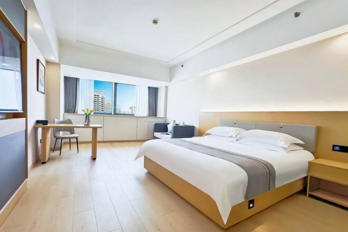 A modern hotel room with a large bed, a desk, a chair, and a view of the cityscape through the window.
