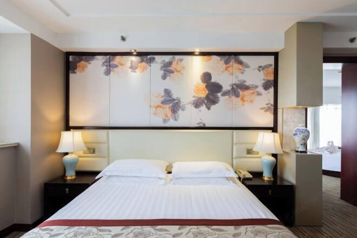 A hotel room with a bed, two bedside tables with lamps, a large floral artwork on the wall, and a nightstand with a vase.