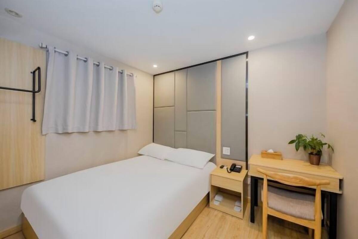 A modern hotel room with a bed, a desk, a chair, a mirror, and a potted plant.