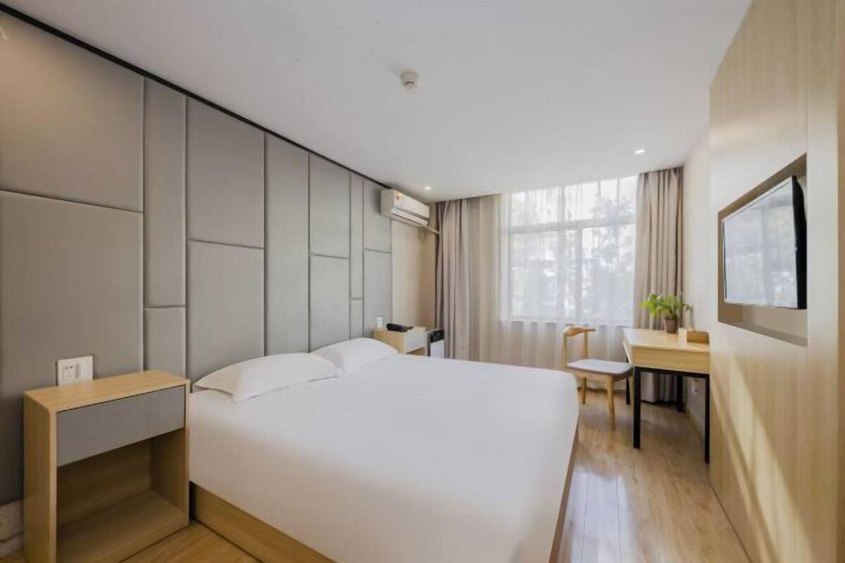 A modern hotel room with a large bed, a desk, and a chair, a TV mounted on the wall, and a window with curtains.
