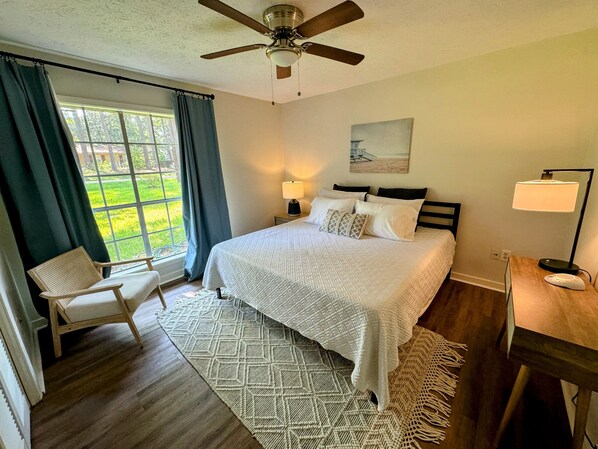 A bedroom with a bed, a chair, a desk, a lamp, a ceiling fan, and a window with curtains.