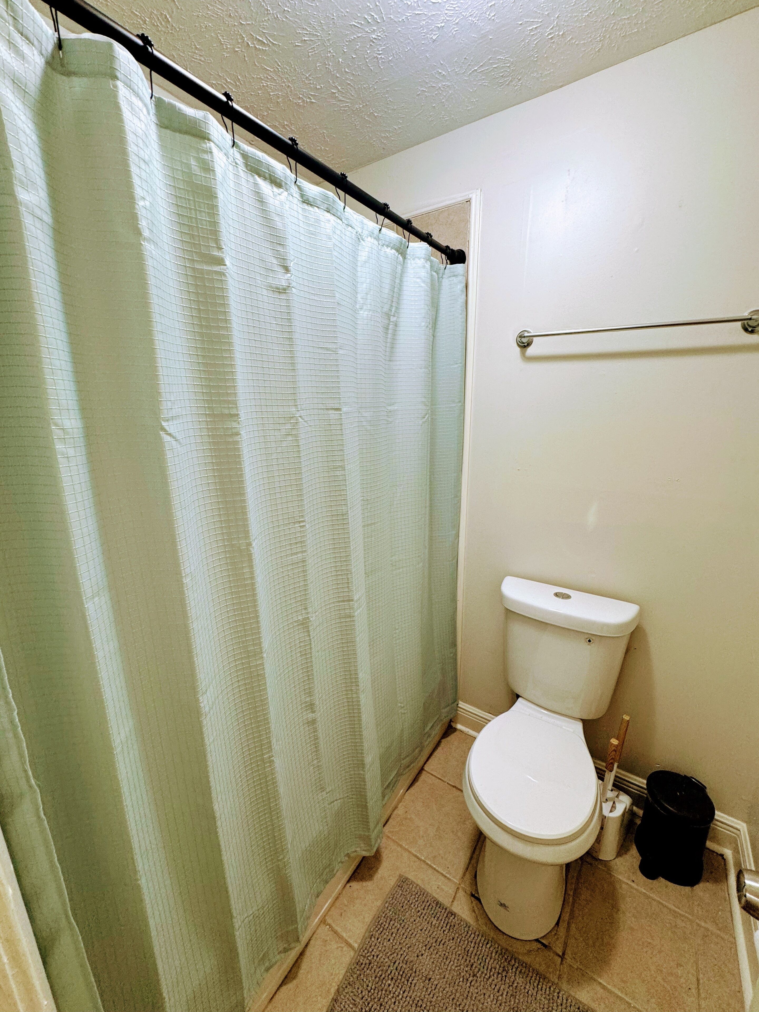 A bathroom with a toilet, a shower curtain, a grab bar, and a small trash bin.