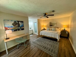 A bedroom with a bed, bedside lamps, a desk, a chair, a ceiling fan, and a painting on the wall.