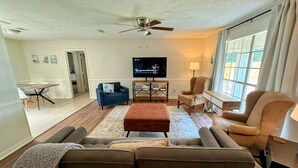 Ein Wohnzimmer mit einer Couch, einem Hocker, Sesseln, einem Fernseher und einem Deckenventilator.