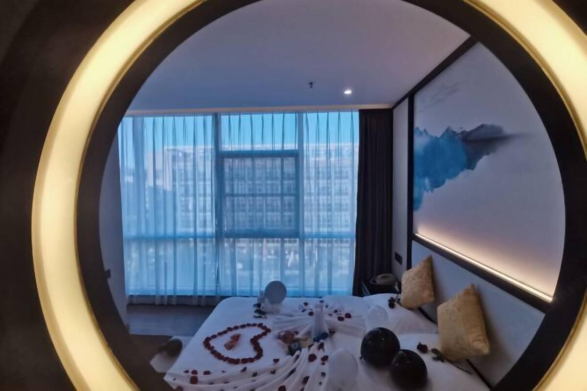 Moderni hotellihuone, jossa on suuri sänky, näkymä kaupunkiin läpikuultavien verhojen läpi ja taulu seinällä.