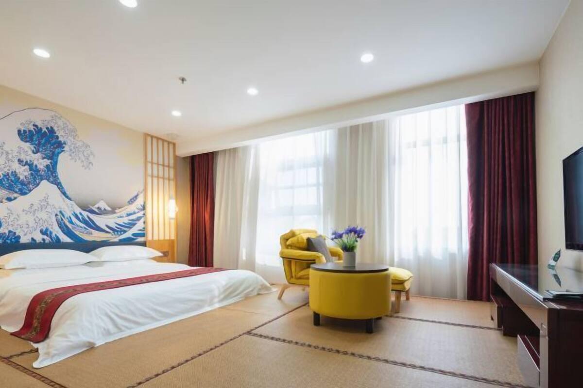 Hotellihuone, jossa on suuri sänky, keltainen nojatuoli, pieni pyöreä pöytä maljakolla ja jalustalla seisova televisio.