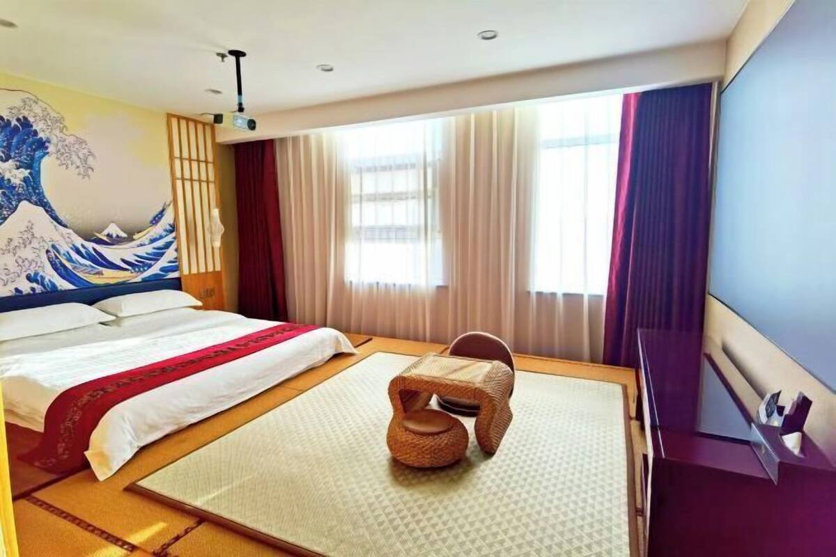 Hotellihuone, jossa on sänky, tatamimatto, punottu tuoli ja suuri ikkuna, jossa on verhot.