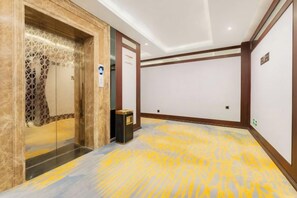 A hotel corridor with a patterned carpet, a glass elevator, and a door with a decorative design.