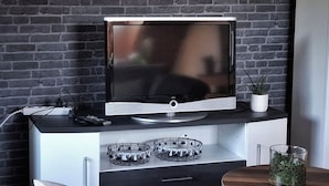 A modern living room with a flat-screen TV on a black and white cabinet, a small potted plant, and a glass container on a wooden table.