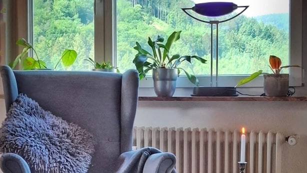 A modern living room with a grey armchair, a matching ottoman, a small round table with a candle, and a view of a forest through large windows.