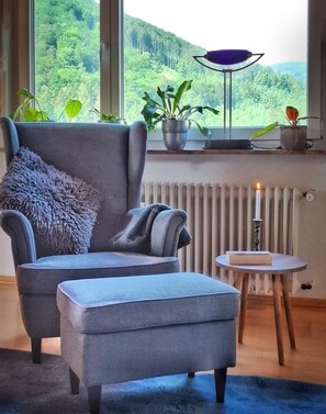 A modern living room with a grey armchair, a matching ottoman, a small round table with a candle, and a view of a forest through large windows.