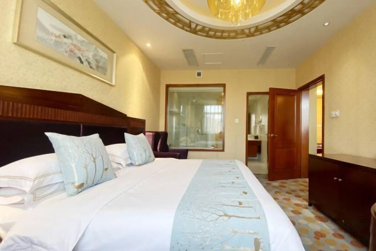 Een hotelkamer met een groot bed, een houten hoofdeinde, een kroonluchter, een ingelijst schilderij aan de muur en een deur naar een andere kamer.