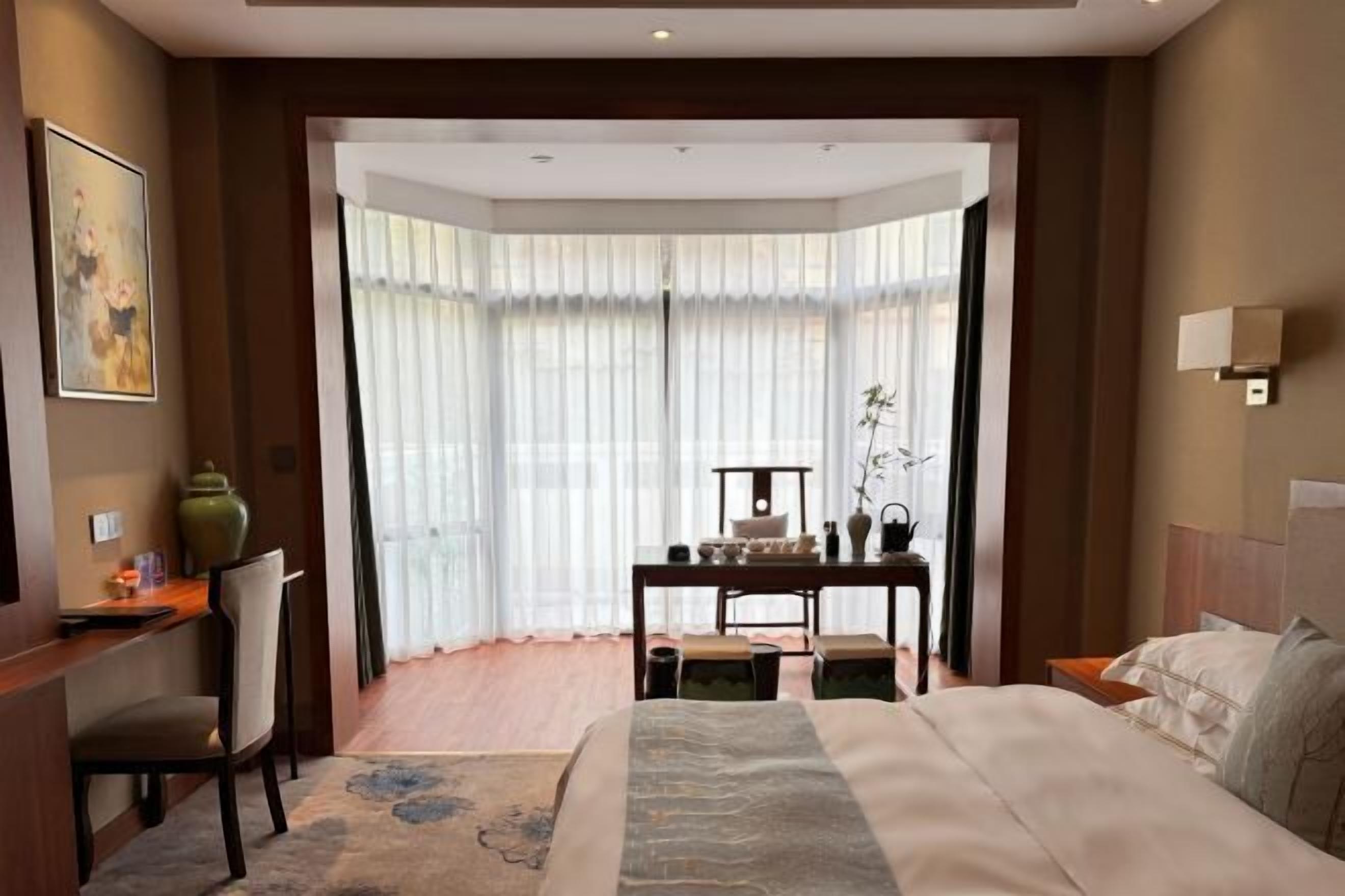 A hotel room with a large bed, a desk with a chair, a small table with chairs, and a large window with sheer curtains.