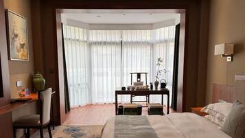 A hotel room with a large bed, a desk with a chair, a small table with chairs, and a large window with sheer curtains.