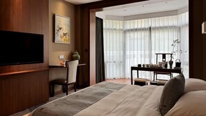A hotel room with a large bed, a flat-screen TV mounted on the wall, a desk with a chair, and a view of the outdoors through sheer curtains. - Kaiyuan Hot Spring Resort (Dezhou)