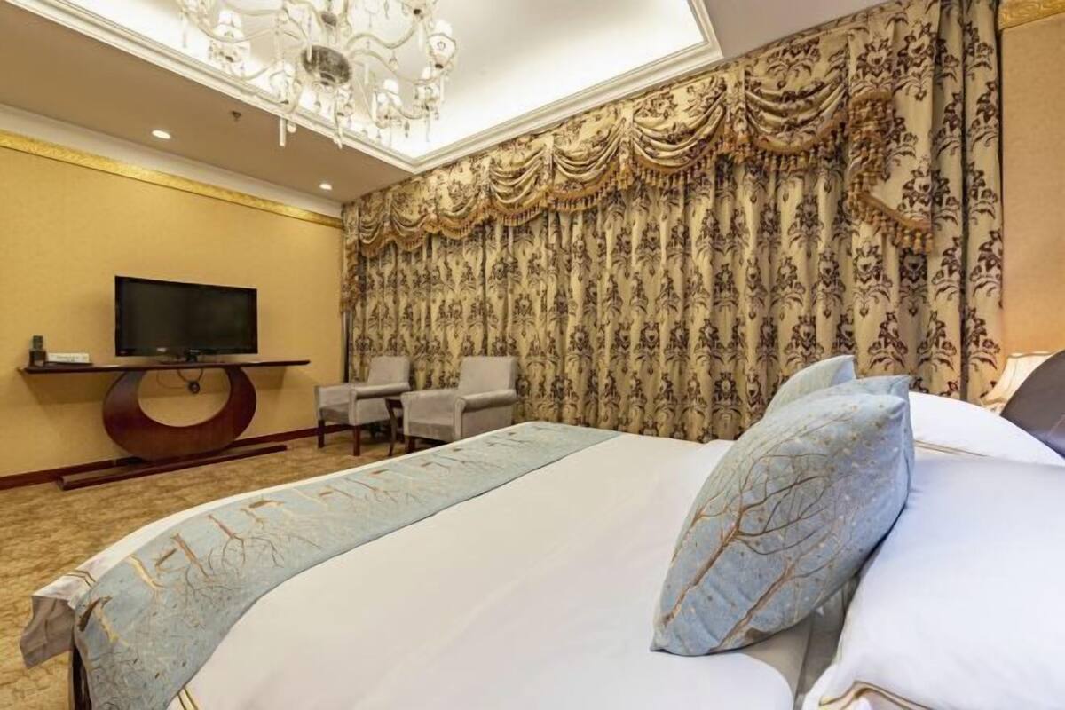 Een hotelkamer met een groot bed, een televisie op een standaard, een kroonluchter en decoratieve gordijnen.