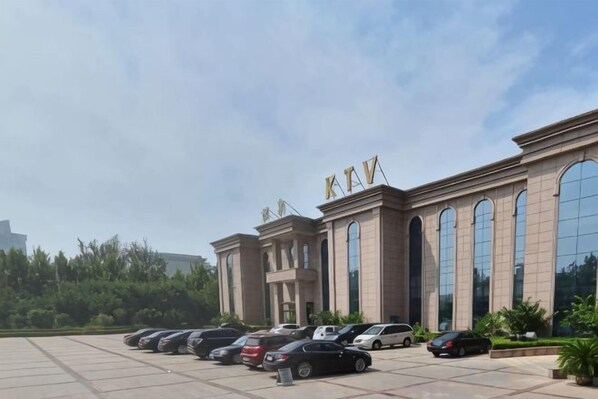 A building with the letters 'KAV' on the facade, a parking lot with several cars, and a clear sky. - Kaiyuan Hot Spring Resort (Dezhou)