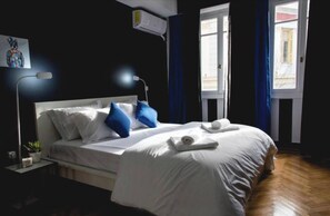 A neatly made bed with white linens and blue pillows, a bedside lamp, and a small table with a plant.