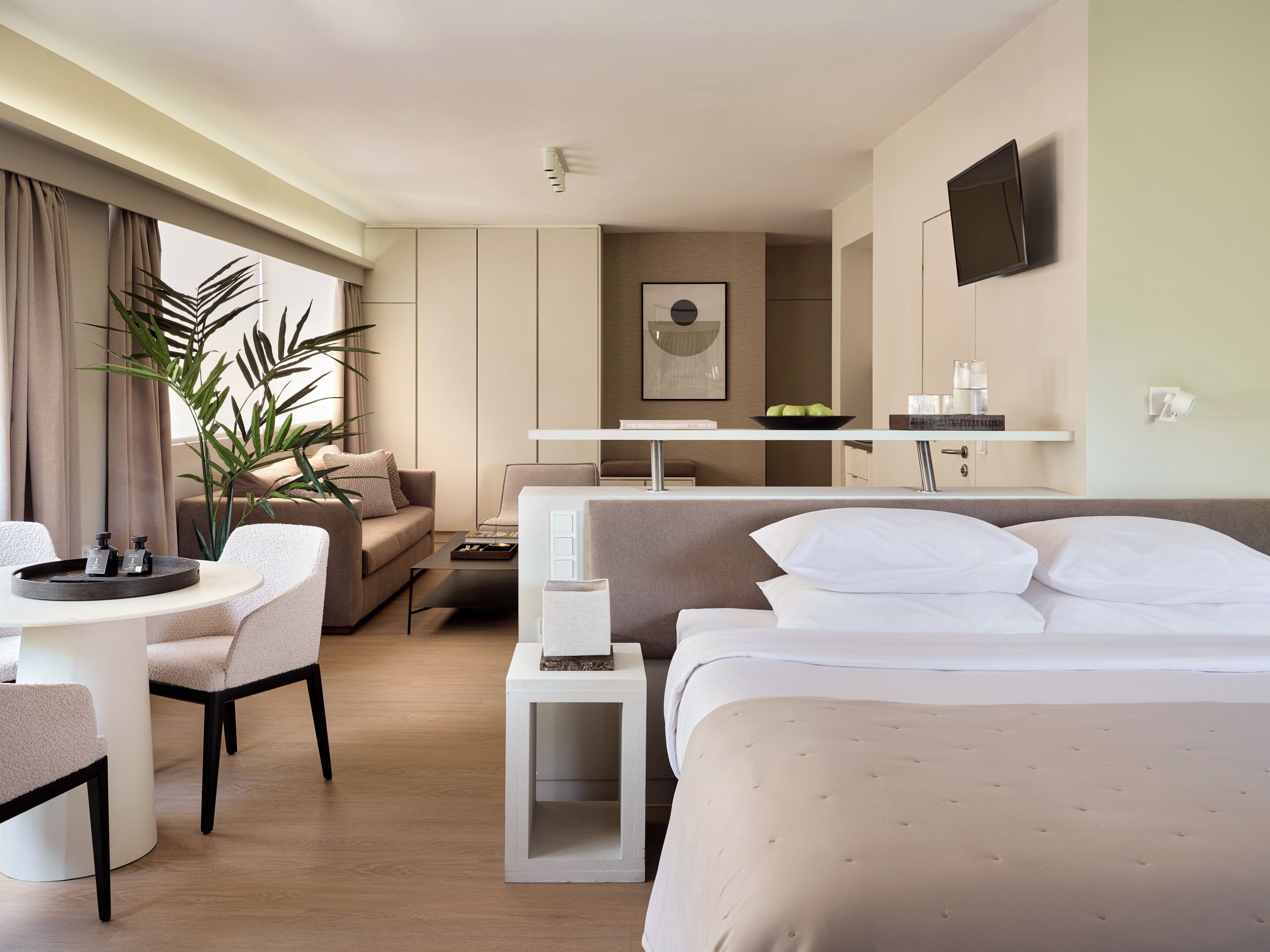 A modern hotel room with a bed, a sofa, a dining table, a small side table, a TV, and a potted plant.