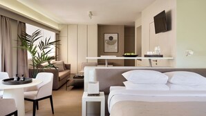 A modern hotel room with a bed, a sofa, a dining table, a small side table, a TV, and a potted plant.