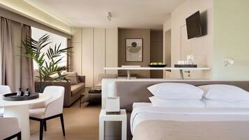 A modern hotel room with a bed, a sofa, a dining table, a small side table, a TV, and a potted plant.