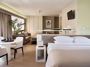 Una habitación de hotel moderna con cama, sofá, mesa de comedor, mesita auxiliar, televisor y planta en maceta.