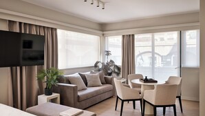 A modern hotel room with a flat-screen TV, a sofa, a dining area with chairs, a small table, and a potted plant. - Momo Suites Acropolis by Aura Homes (Athens)