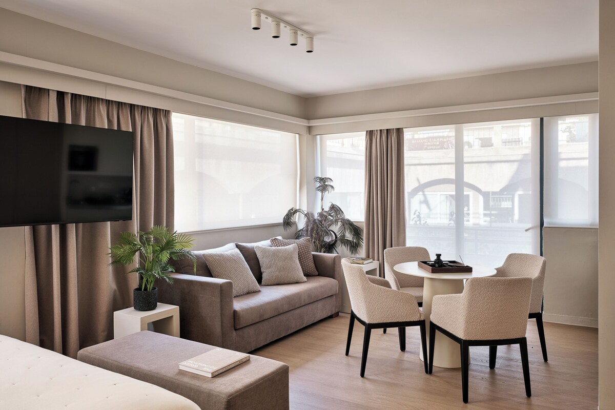 Una habitación de hotel moderna con una televisión de pantalla plana, un sofá, una zona de comedor con sillas, una pequeña mesa y una planta en maceta.