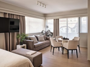 A modern hotel room with a flat-screen TV, a sofa, a dining area with chairs, a small table, and a potted plant. - Momo Suites Acropolis by Aura Homes (Athens)