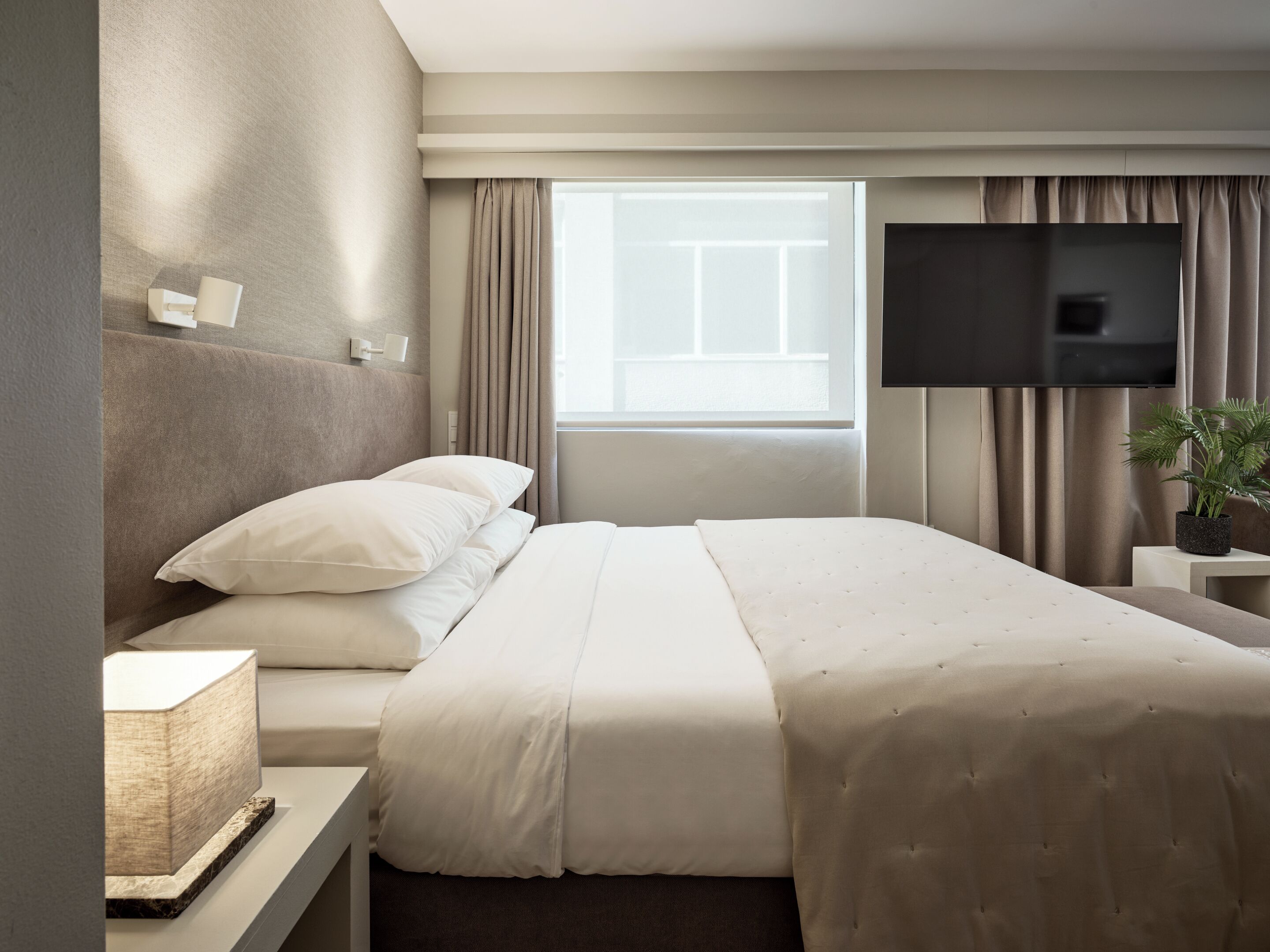 A hotel room with a bed, a flat-screen TV, a bedside table, a lamp, and a window with curtains.