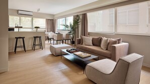 A modern living room with a sofa, ottoman, and coffee table. There is a dining area with chairs and a table, and a kitchen area with a bar stool. - Momo Suites Acropolis by Aura Homes (Athens)
