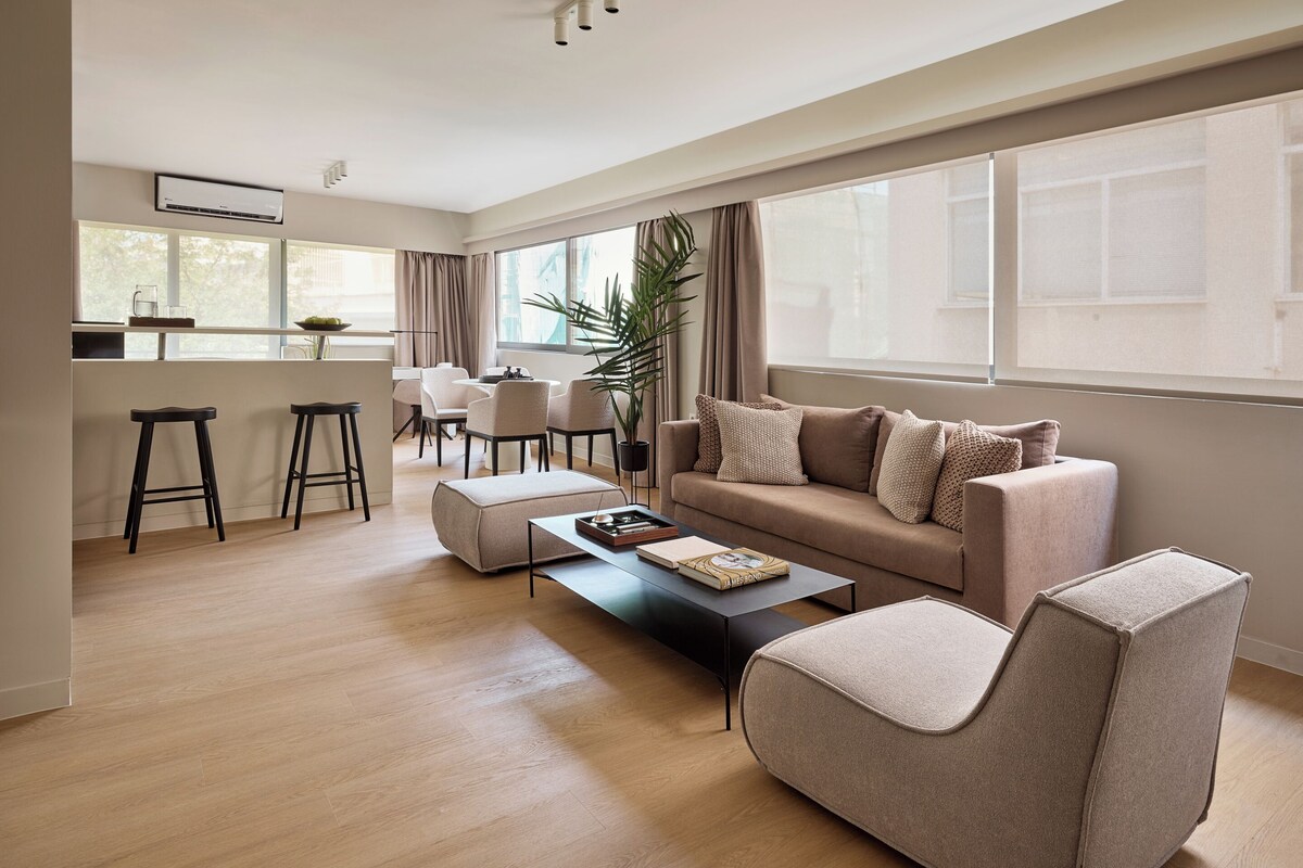 Una sala moderna con sofá, reposapiés y mesa de centro. Hay un comedor con sillas y una mesa, y una cocina con taburete de bar.
