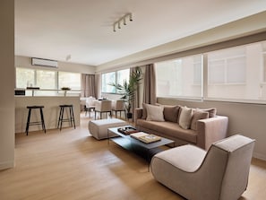 A modern living room with a sofa, ottoman, and coffee table. There is a dining area with chairs and a table, and a kitchen area with a bar stool. - Momo Suites Acropolis by Aura Homes (Athens)