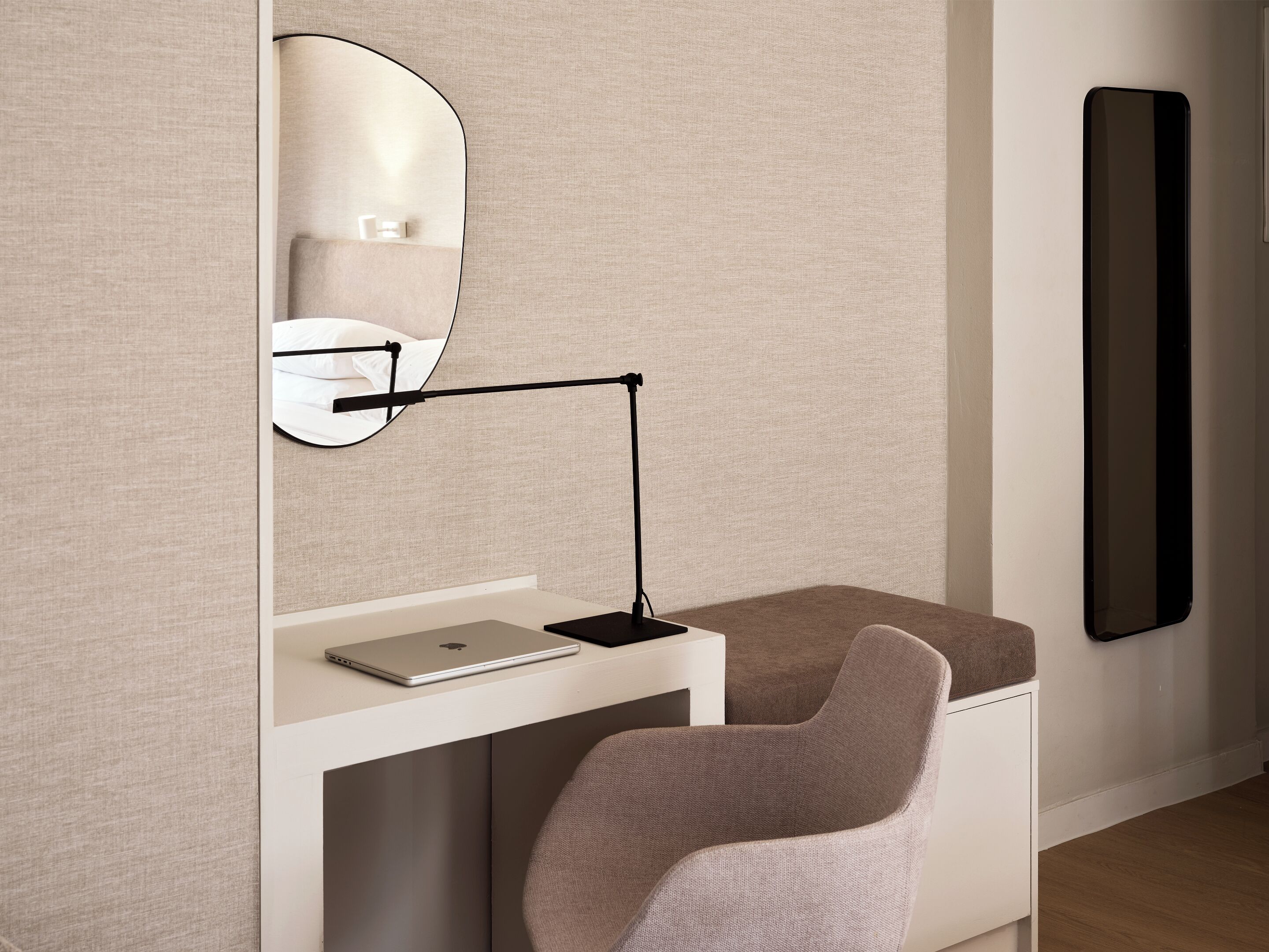 A modern room with a white desk, a laptop, a chair, a wall-mounted mirror, and a large mirror.
